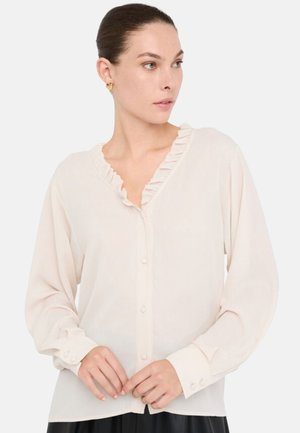 Blusa beige chiaro, con scollo arricciato, maniche lunghe, chiusura con bottoni, tessuto morbido e dettagli sui polsini; risultante in uno stile con un lieve lucido.