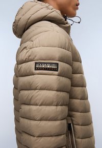 Giacca puffer beige con cappuccio, caratterizzata da sezioni trapuntate, un'etichetta nera sul braccio e una tasca laterale con dettaglio zip.