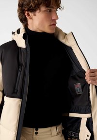 Tweekleurige gewatteerde jas met een zwarte bovenkant en beige onderkant, voorzien van ritsen, logo-accenten en een binnenzak. Gladde textuur.