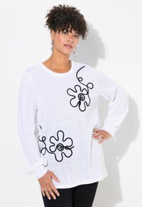 Vit stickad tröja med svart blommönster och böjda linjer. Långa ärmar med ribbade manschetter. Bärs över svarta byxor.