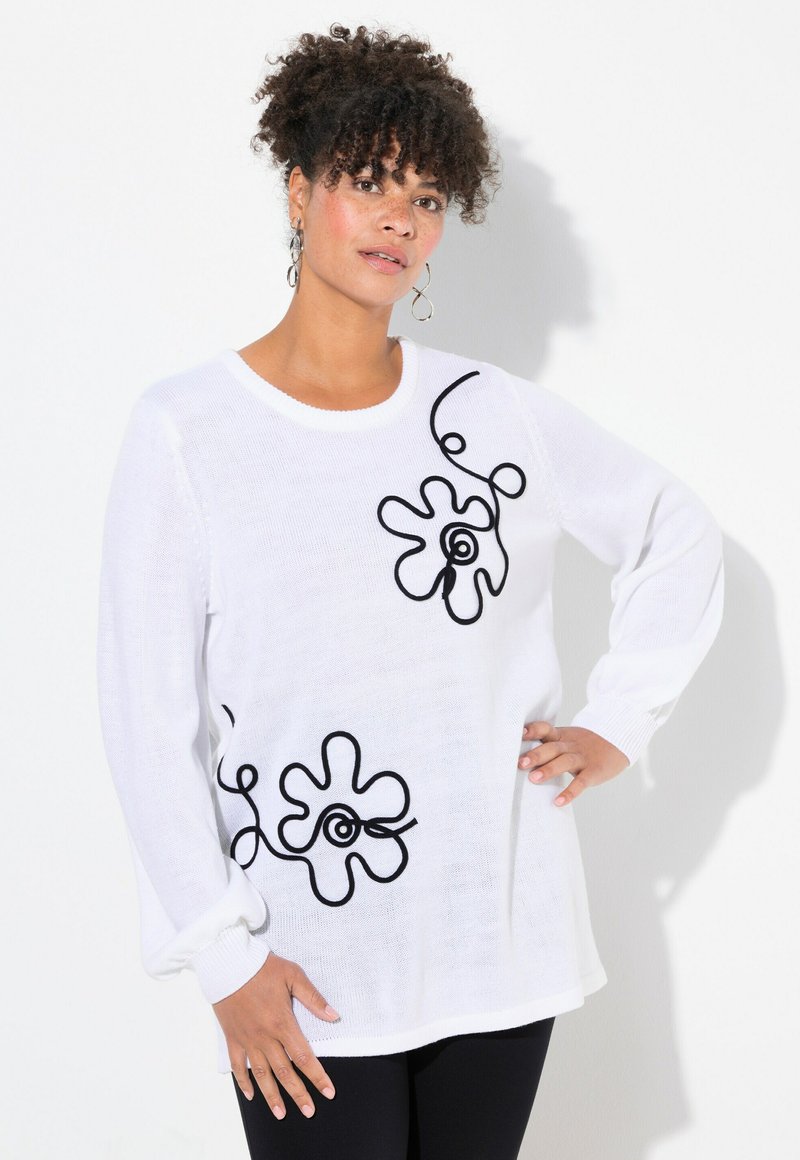 Vit stickad tröja med svart blommönster och böjda linjer. Långa ärmar med ribbade manschetter. Bärs över svarta byxor.