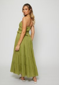 Woman with long blonde hair in a strapless olive green tiered maxi dress and white sandals, looking over her shoulder against a plain background.