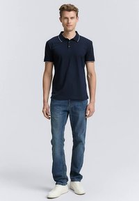 Tamsiai mėlynas trumpomis rankovėmis polo marškinėliai su baltais apykaklės akcentais, derinami su šviesiai mėlynais džinsais ir baltais sportiniais bateliais. Lengvos medžiagos, klasikinio kirpimo.