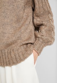 Paksu neulepaita vaaleanruskeana, jossa on resorireunus ja palmikkoneulekuvio hihansuissa, yhdistetty valkoisiin housuihin.