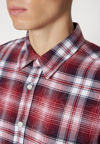 Gros plan d'une personne portant une chemise à carreaux rouge, blanche et bleu marine à boutons avec un col pointu, montrant la partie inférieure du visage et le cou.