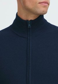 Uspravljen prikaz donjeg dijela lica i vrata muškarca koji nosi tamnoplavi duksericu s rajsferšlusom, s rebrastim visokim ovratnikom zatvorenim do vrha.