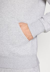 Ruka spočívajúca vo vrecku svetlosivej mikiny s rebrovaným lemom na manžete a pásiku.