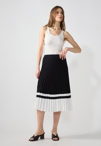 Zwart-witte plooienrok met een zwarte tailleband, met contrasterende horizontale strepen. Model draagt een witte tanktop en zwarte hakken.