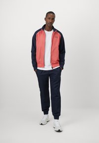 Veste de survêtement en corail et marine, avec fermeture éclair, deux poches avant et poignets en côte ; associée à un pantalon de jogging navy et des baskets blanches.