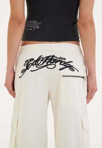Zwarte cropped tanktop met logo aan de zijkant, gecombineerd met crèmekleurige cargobroeken met een zwart geborduurd ontwerp en een zijritszak.