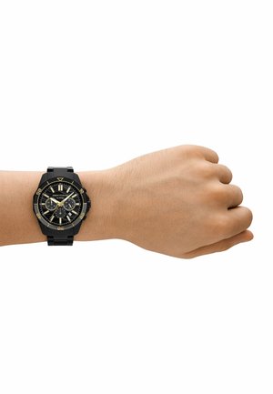 Černé analogové náramkové hodinky se zlatými akcenty, s kulatým ciferníkem, chronografovými ukazateli a robustním náramkem na světlejším tónu pleti.