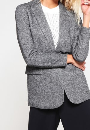 Woman wearing grey textured blazer over white dotted shirt with arms crossed, shown from neck to hips.