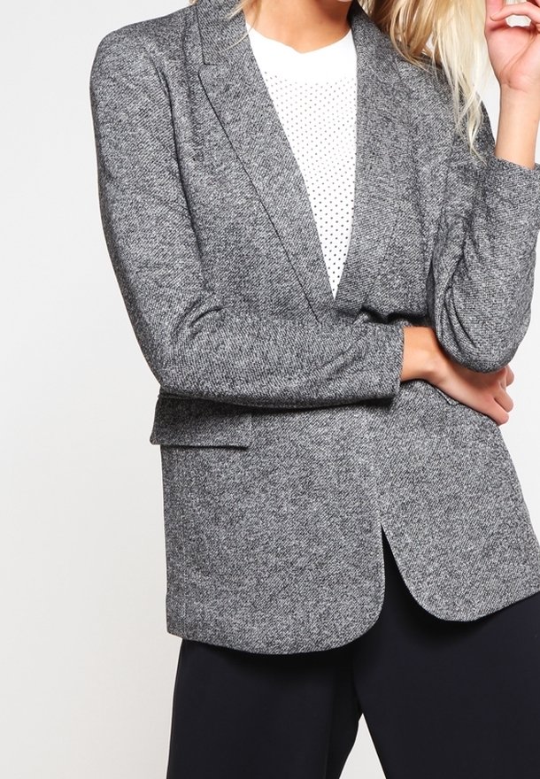 Femme portant un blazer gris texturé sur une chemise blanche à pois, les bras croisés, vue du cou aux hanches.