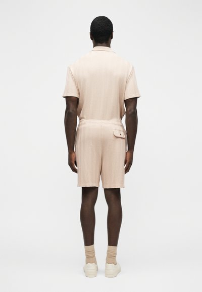 Homme debout de dos, portant une chemise à manches courtes beige clair et un short assorti avec une poche arrière boutonnée, des chaussettes beiges et des chaussures blanches.