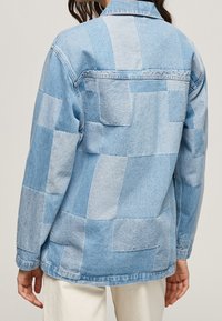 Denim takki, jossa on erikoispainatus eri sinisen sävyissä, kaulus, pitkät hihat ja kaksi takataskua.