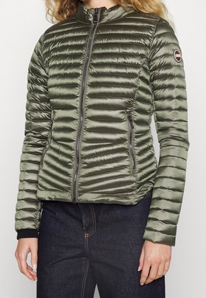 Mujer con chaqueta ajustada acolchada de color verde oliva con cremallera frontal y bolsillos laterales con cremallera, combinada con vaqueros azul oscuro.