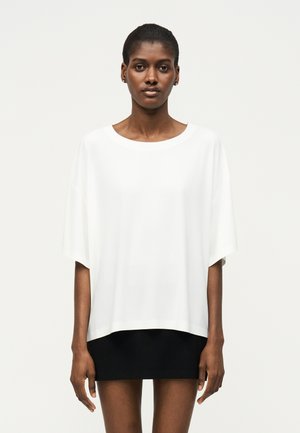 Γυναίκα με κοντά μαλλιά, που φοράει άνετη λευκή μπλούζα με κοντά μανίκια συνδυασμένη με μαύρη μίνι φούστα, στέκεται μπροστά σε απλό φωτεινό φόντο.