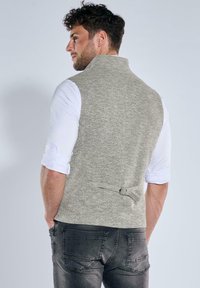 Graue Strickweste mit hohem Kragen und Schnallendetail auf der Rückseite, getragen über einem weißen Langarmshirt, kombiniert mit dunkelgrauen Jeans.