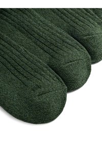 Sötétzöld kötött zoknik bordázott textúrával és megerősített orr résszel. Három pár kiállítva, amelyek egységes dizájnt és színt mutatnak.