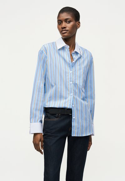 Γυναίκα που φοράει μπλε και λευκό κάθετα ριγέ πουκάμισο με λευκό γιακά και μανσέτες, συνδυασμένο με σκούρο μπλε τζιν και μαύρη ζώνη.