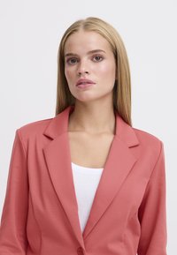 Koraalblazer met een notch-revers, enkele knoopsluiting en lichte stof, gecombineerd met een witte top eronder.