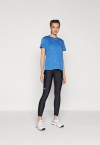Camiseta deportiva azul de manga corta con cuello redondeado, combinada con leggings negros que tienen un logo acentuado y zapatillas deportivas blancas.