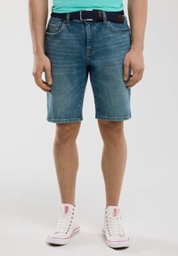 Gaiši zilie džinsi šorti ar taisnu piegriezumu un saplēstu malumu. Saskaņoti ar austu tumši zilu jostu un baltiem augstiem sporta apaviem.