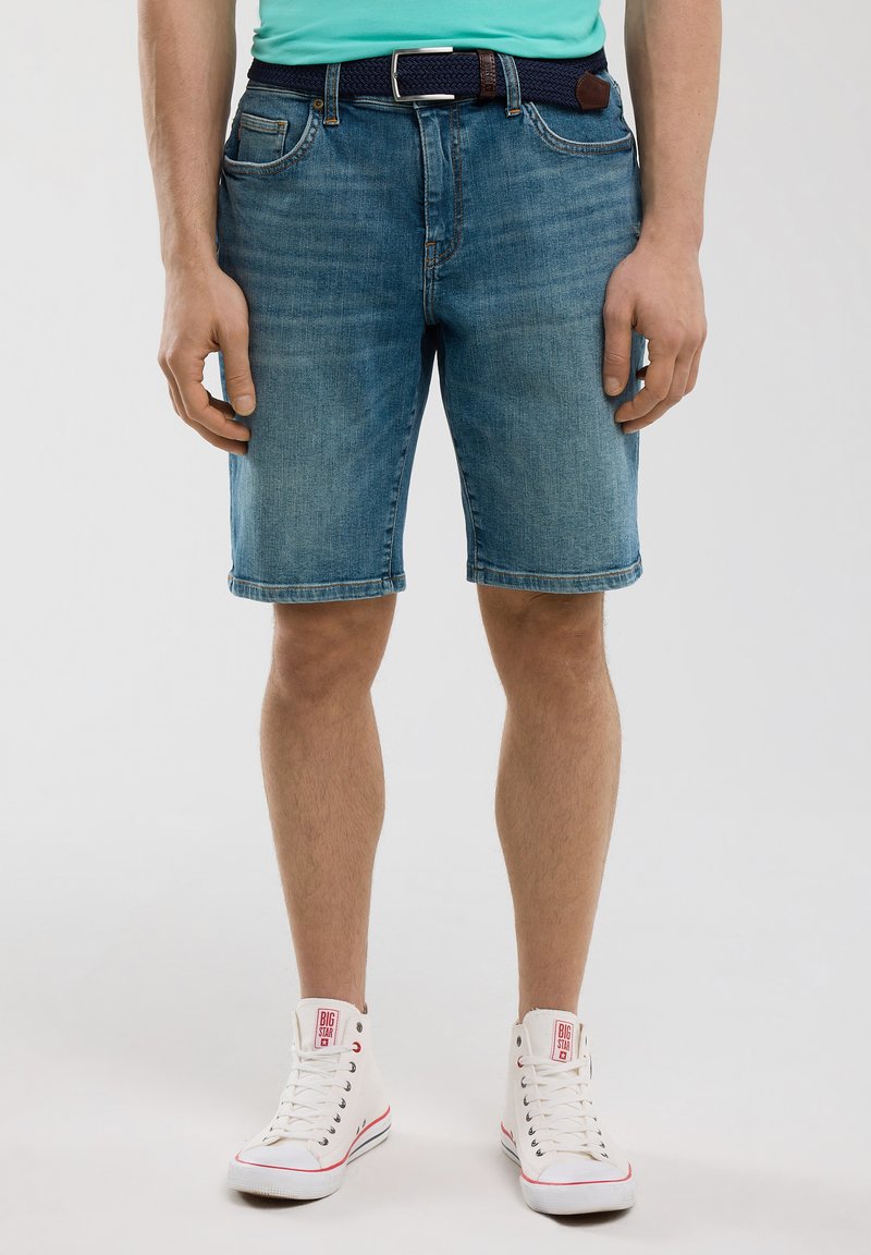 Gaiši zilie džinsi šorti ar taisnu piegriezumu un saplēstu malumu. Saskaņoti ar austu tumši zilu jostu un baltiem augstiem sporta apaviem.