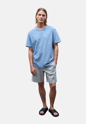 Jeune homme aux cheveux mi-longs portant un t-shirt bleu clair, un short gris et des sandales noires, debout avec une main dans la poche.