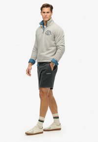 Grijze fleece pullover met een kwart rits en geborduurd logo, gecombineerd met zwarte shorts. Voorzien van blauwe denim manchetten en witte sneakers met bruine zolen.