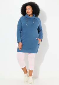 Robe pull bleu avec un col en cowl, présentant des cordons de serrage et des poches latérales, associée à des leggings capri rose clair et des baskets blanches.