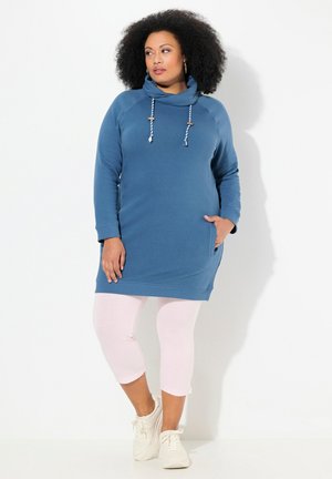 Abito felpa blu con collo avvolgente, dotato di cordini e tasche laterali, abbinato a leggings capri rosa chiaro e sneakers bianche.