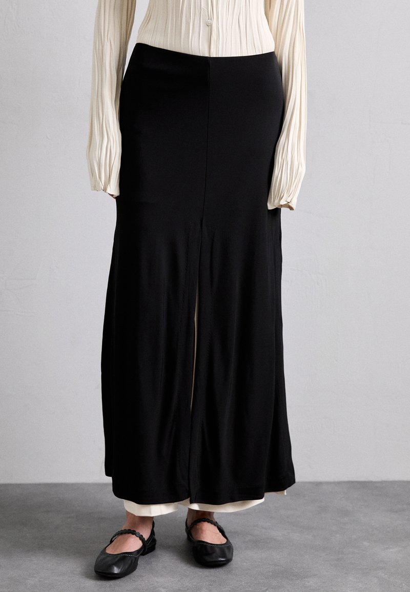 Black maxi skirt made of smooth fabric, featuring a front slit. Paired with a textured light-colored blouse and black flat shoes.