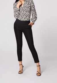 Kvinna i svarta smala byxor, svarta klackskor med remmar och en långärmad blommig skjorta med knappar, stående med en hand i fickan.