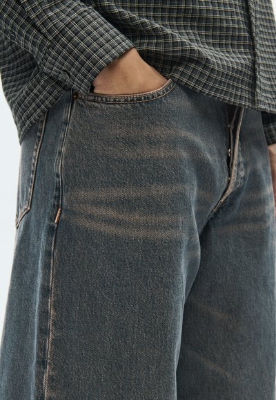 Σκούρο μπλε τζιν με στενή εφαρμογή, με αντίθετα ράμματα και τσέπες. Το υφασμάτινο ύφασμα παρουσιάζει διακριτική φθορά και ραβδώσεις.