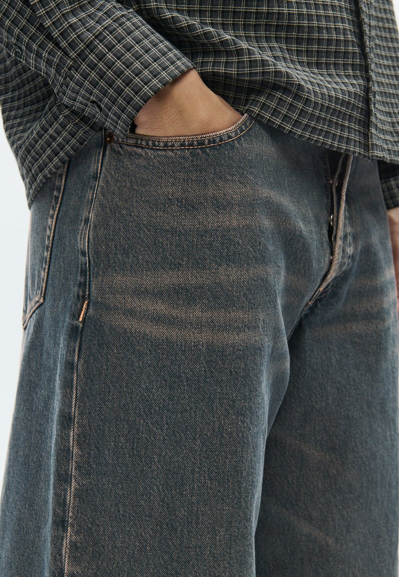 Dark blue denim jeans with a slim fit, featuring contrasting stitching and pockets. Textured fabric shows subtle fading and whiskering.