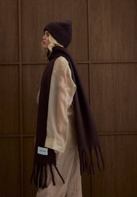 Écharpe en laine tricotée de couleur brun foncé avec de longues franges, assortie à un bonnet assorti, portée sur un chemisier léger en soie, sur un fond de panneaux en bois.