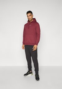 Bordeaux hættejakke med frontlomme og snore, parret med sorte bukser og sorte sneakers med grønne accenter. Ensfarvet baggrund.
