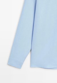 Light blue long-sleeve shirt with a soft texture, rounded hem, cuff finish, and subtle stitching details. Casual, relaxed fit.