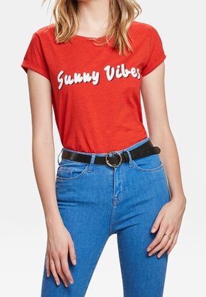 Kobieta ubrana w czerwoną koszulkę z napisem „Sunny Vibes” wpuszczoną w wysokowodne niebieskie dżinsy z czarnym paskiem i okrągłą klamrą.