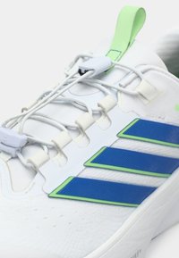 Chaussure de sport blanche avec des accents bleus et verts, présentant une tige synthétique texturée, des lacets élastiques et une languette pour un enfilage facile.
