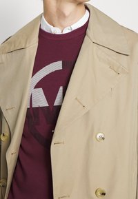Abrigo beige tipo trench con diseño de doble botonadura, grandes botones y cuello con muesca, superpuesto a una sudadera burdeos con un atrevido estampado gráfico.