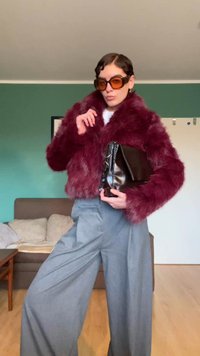 Άτομο που φοράει μπουφάν από βελούδινη γούνα χρώματος μπουρντό, μεγάλα καφέ πορτοκαλί γυαλιά ηλίου και γκρι παντελόνι με φαρδύ πόδι, κρατώντας μια μαύρη δερμάτινη τσάντα μέσα σε εσωτερικό χώρο.