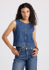 Young woman with curly hair wearing sleeveless denim top and matching denim jeans, standing with hands near pockets against a plain background.