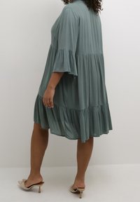 Green, tiered dress with a relaxed fit, featuring flared sleeves and a lightweight, textured fabric. The hemline is slightly ruffled.