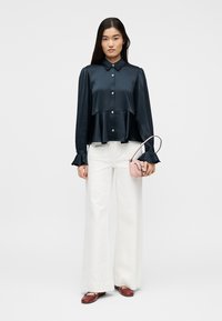 Γυναίκα που φοράει μαύρη μπλούζα με κουμπιά και φουσκωτά μανίκια, άσπρο παντελόνι με φαρδιά μπατζάκια, καφέ παπούτσια και κρατά ένα μικρό ροζ τσαντάκι.