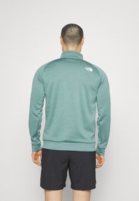 Uomo che indossa una giacca zip-up verde acqua e pantaloni corti neri, in piedi con le spalle rivolte verso un fondo bianco semplice.