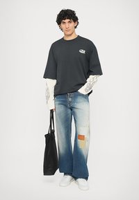 T-shirt oversized nera con logo, sovrapposta a maniche lunghe bianche. Jeans azzurri chiari strappati con toppe. La modella porta una borsa tote nera.