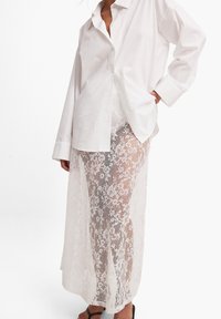 Hvid oversized skjorte med knaplukning parret med en gennemsigtig tyl-maxikjole med blomsterprint og en løs, flagrende silhuet.