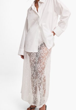 Camicia bianca oversize con bottoni abbinata a una gonna maxi in pizzo trasparente con motivi floreali e una silhouette ampia e fluida.